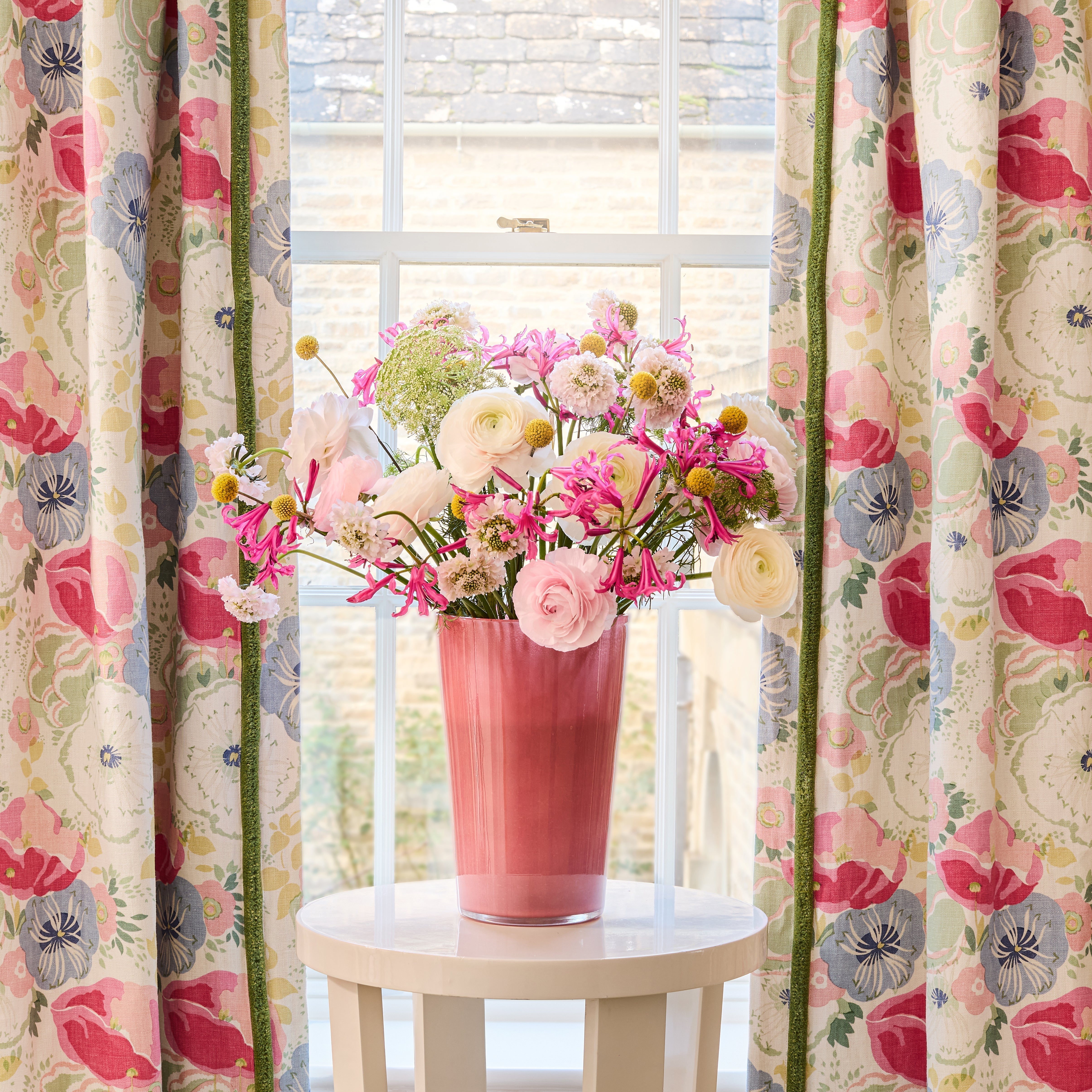 Joni Raspberry floral printed linen curtains by Linwood, featuring pink, blue, and green blooms, framing bright windows with a white table and matching floral arrangement.